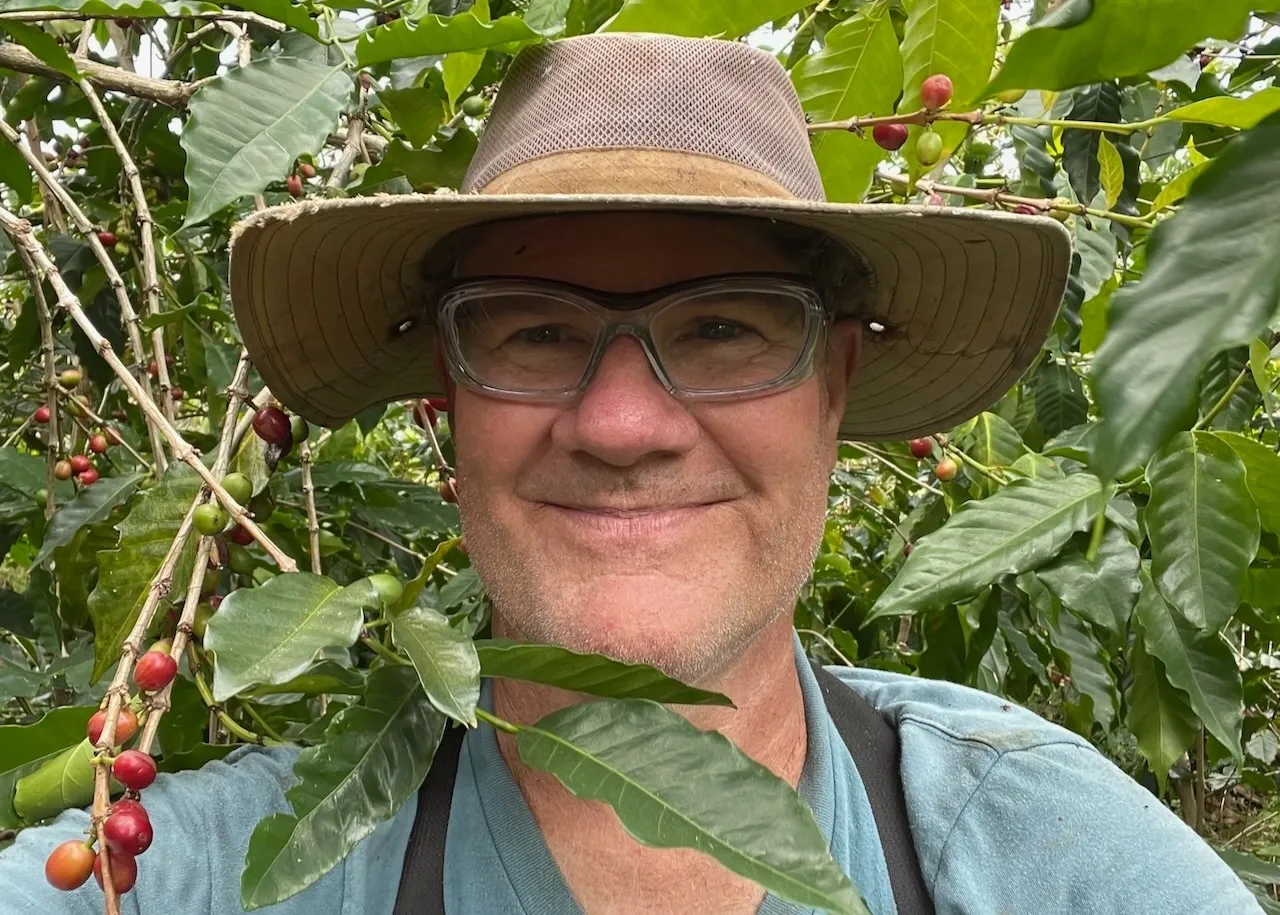 A man wearing glasses and hat standing in front of trees.